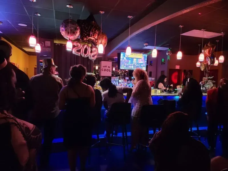 Dim nightclub with people seated at a glowing blue bar under red lights and hanging disco balls.