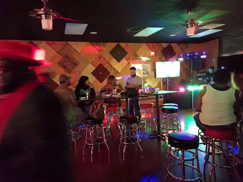 Dimly lit bar with people seated at a high table, neon lights, and a checkered wall backdrop
