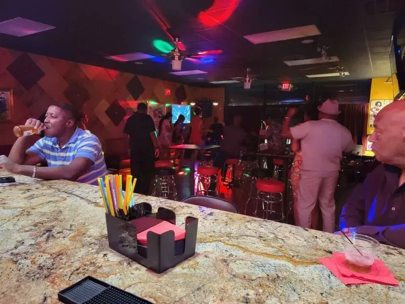 Dimly lit nightclub bar with people seated and standing, colorful neon lights and patterned wall decor.