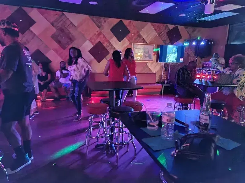 Dim nightclub with people dancing, neon pink-purple lighting, and tables with drinks in the foreground
