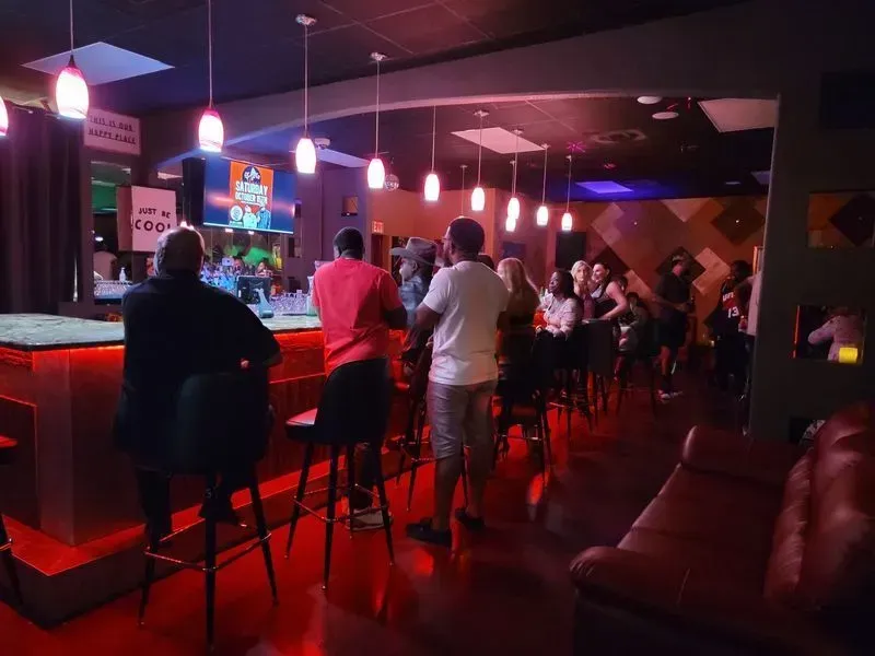 Dimly lit bar with red stools, patrons at the counter, and people gathered in the background