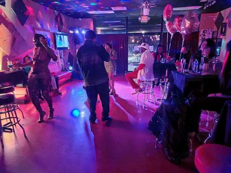 Neon-lit nightclub dance floor with people socializing, pink and blue lights, and balloons overhead