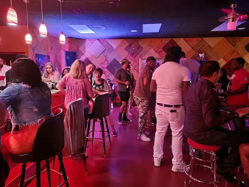 Crowded bar with people seated at tables, pink-purple lighting, and a man in white standing near the center