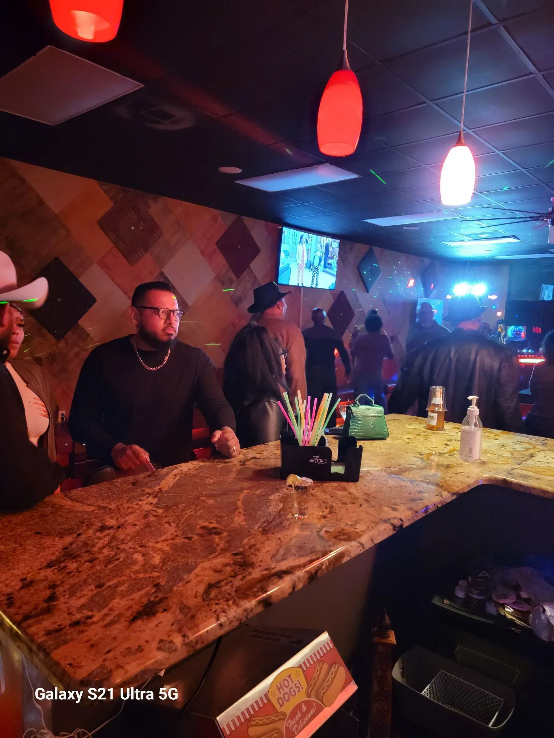 Dimly lit bar with red pendant lights, marble counter, and patrons gathered inside