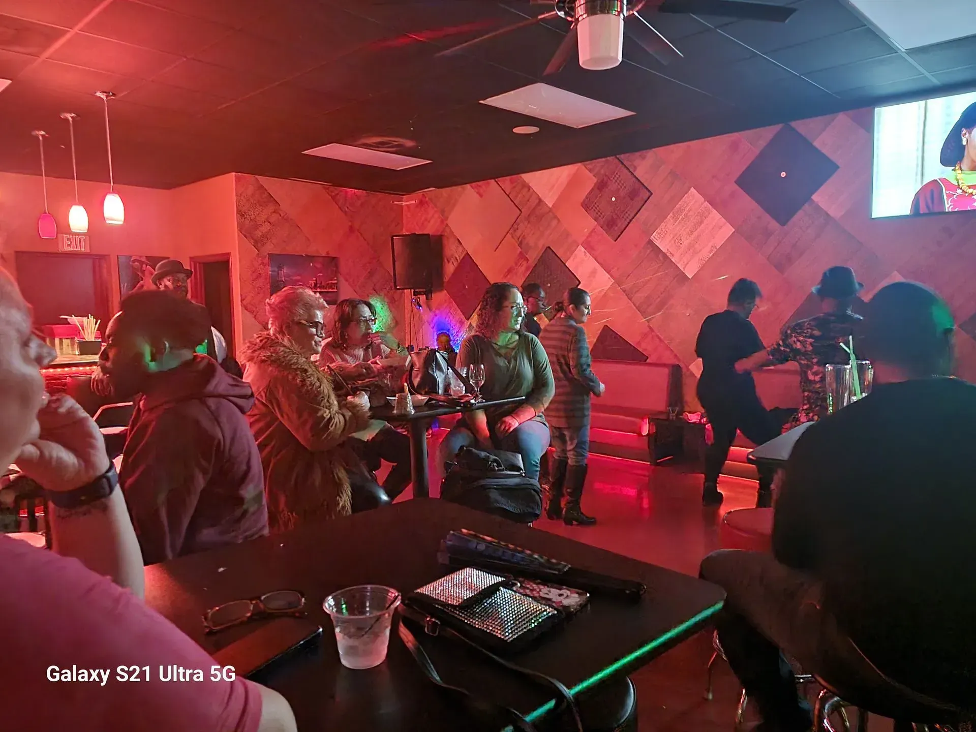 People socializing in a dimly lit lounge with red and green lights, tables, and a wall-mounted TV.