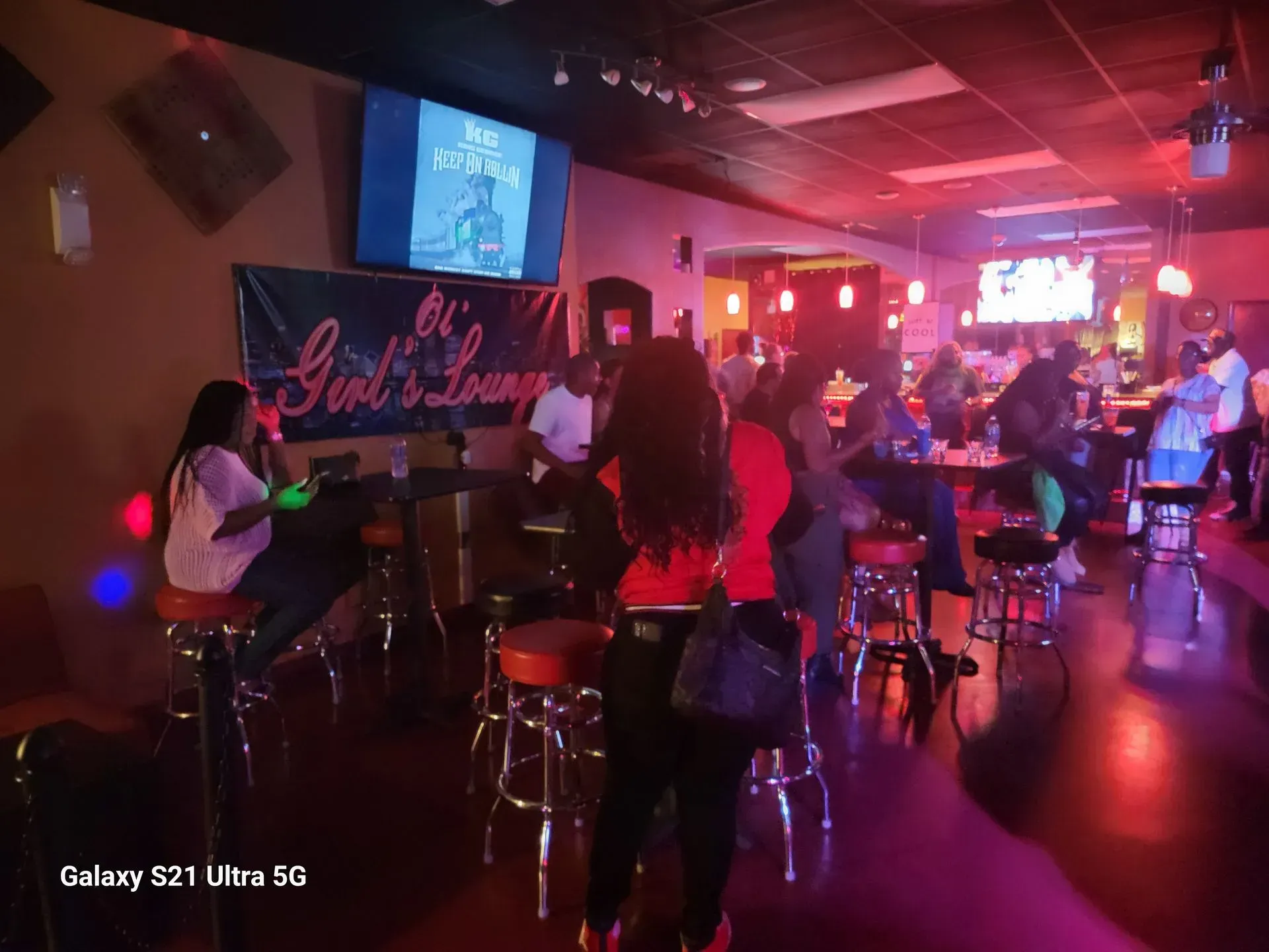 Dimly lit bar with patrons seated at tables, red and purple lights, and a karaoke screen overhead