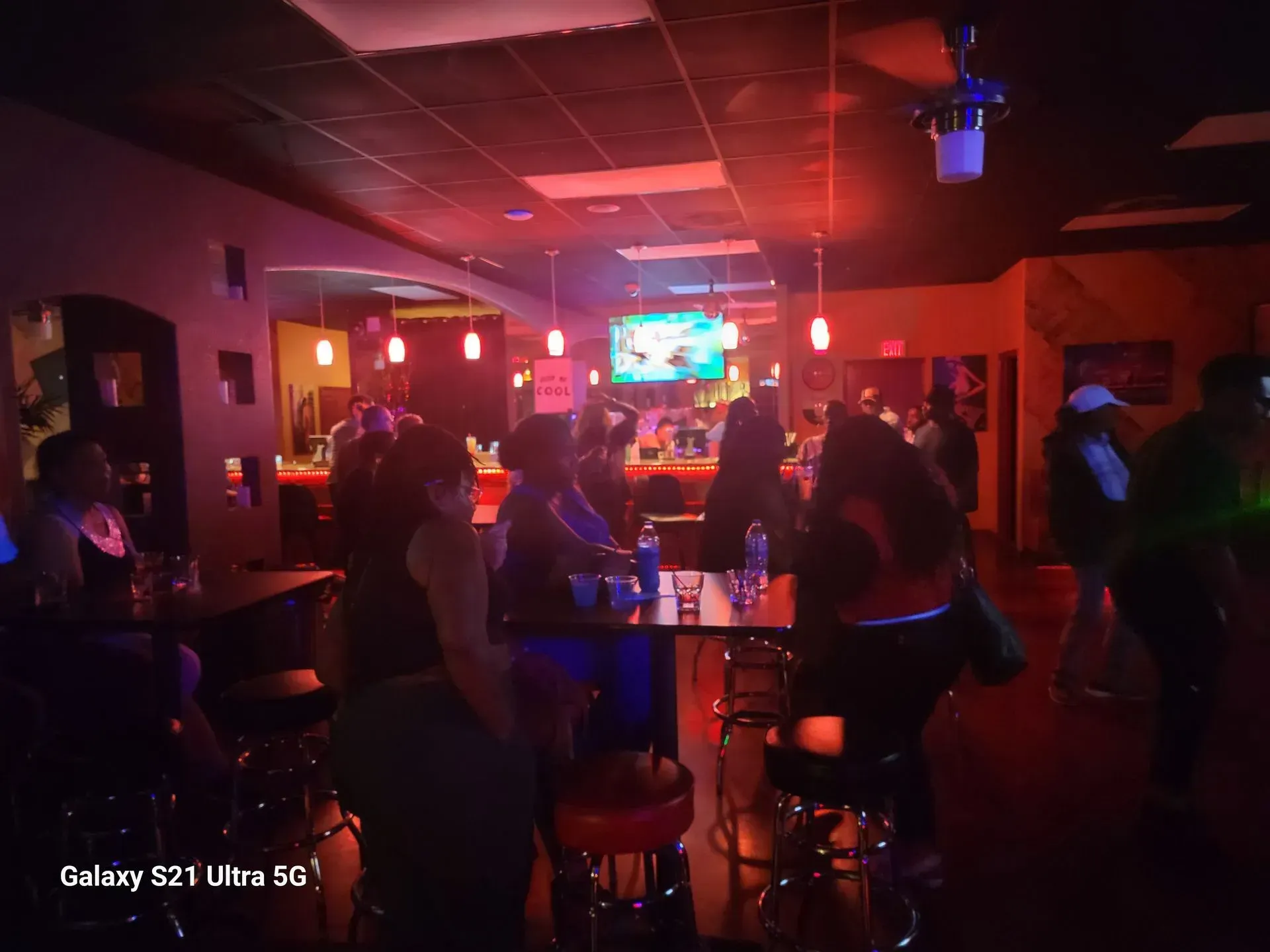Dimly lit bar with people dancing under red and purple lights near a TV screen