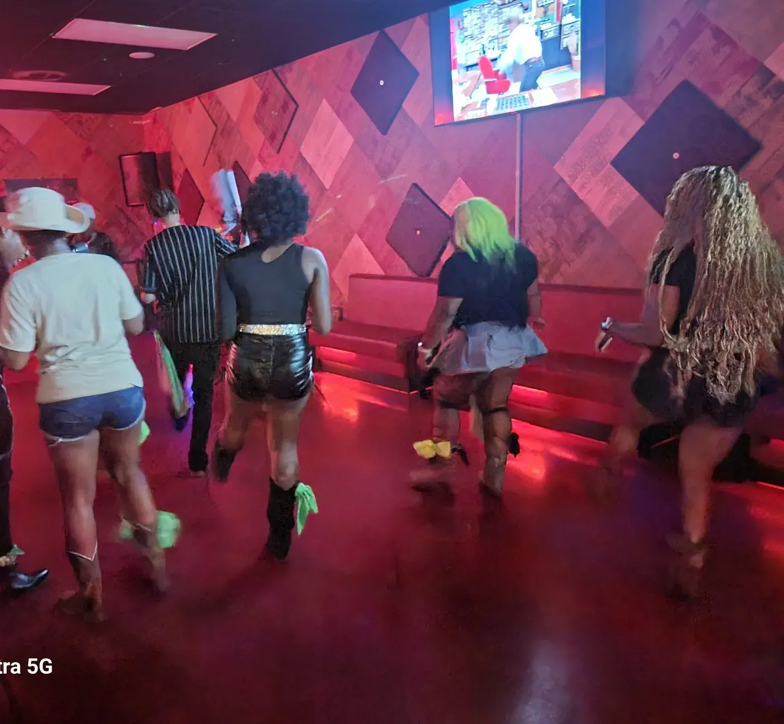 People walking in a neon-lit dance club with pink walls and a TV screen overhead