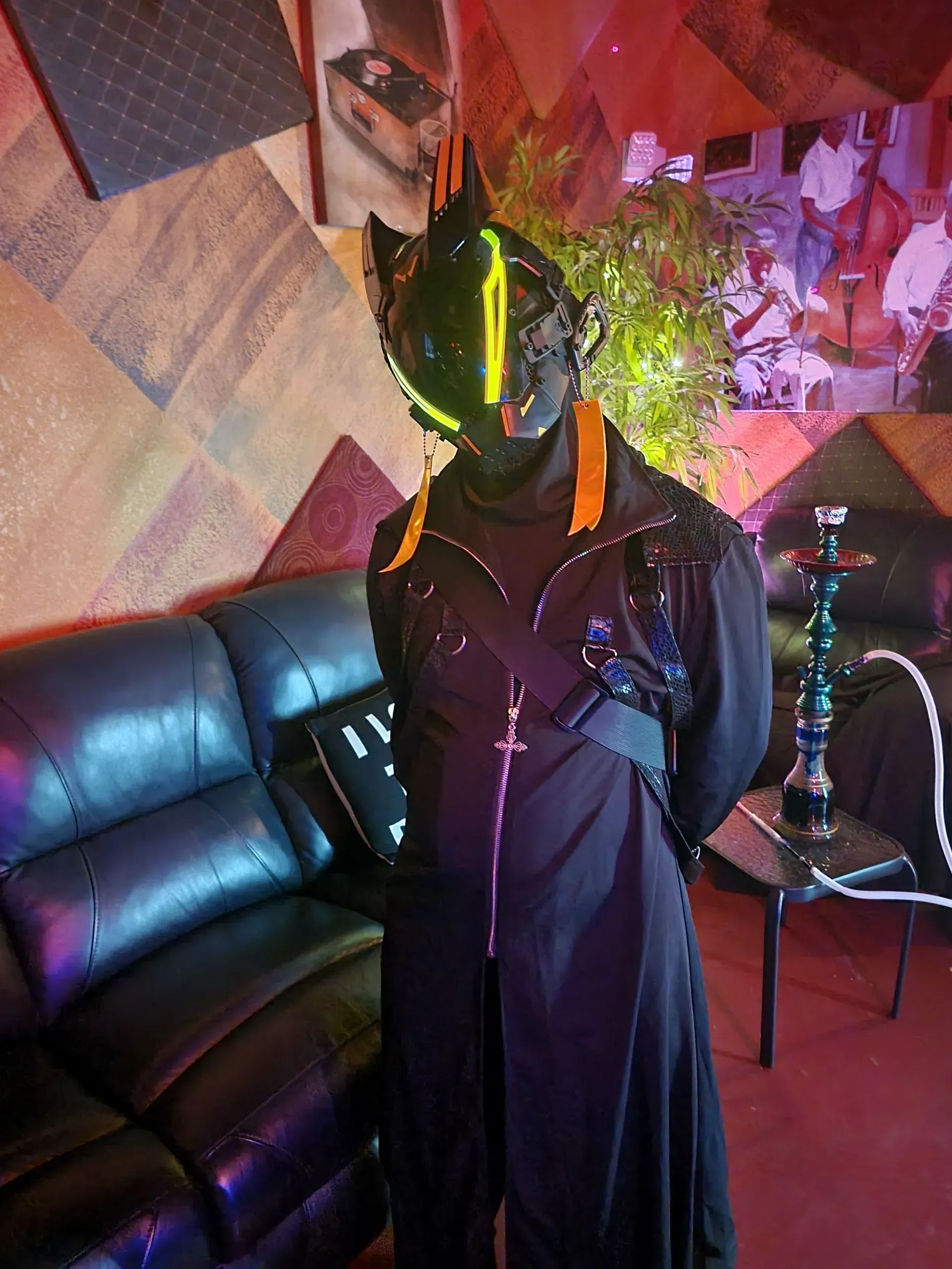 Person in a black outfit and neon helmet standing in a colorful lounge beside a leather couch and hookah.