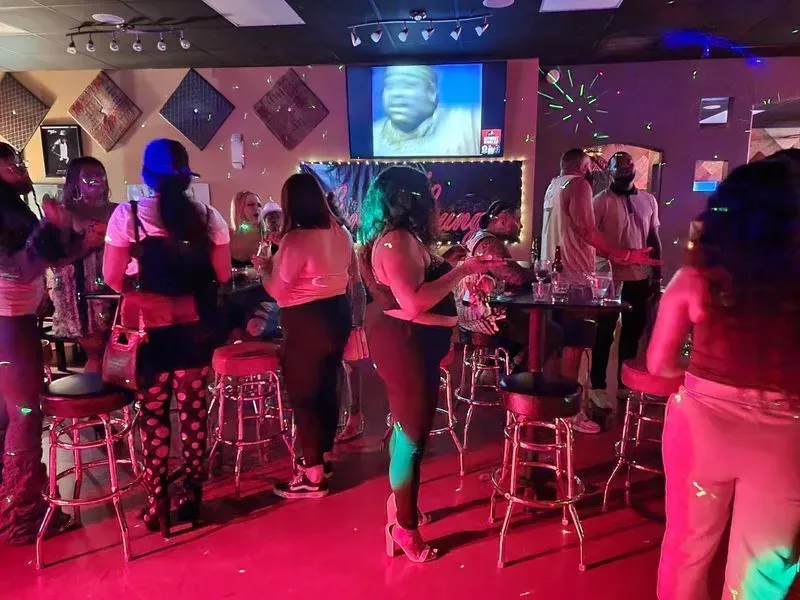 Crowded neon-lit nightclub with people standing at tables and a screen showing a face in the background