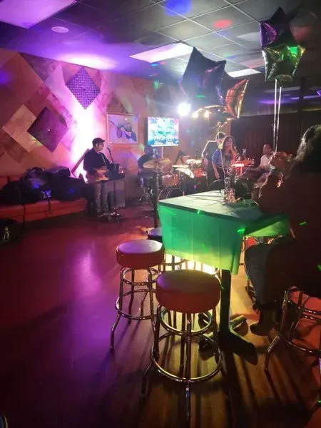 Dimly lit bar with people seated at tables, purple lights, and musicians performing on stage.