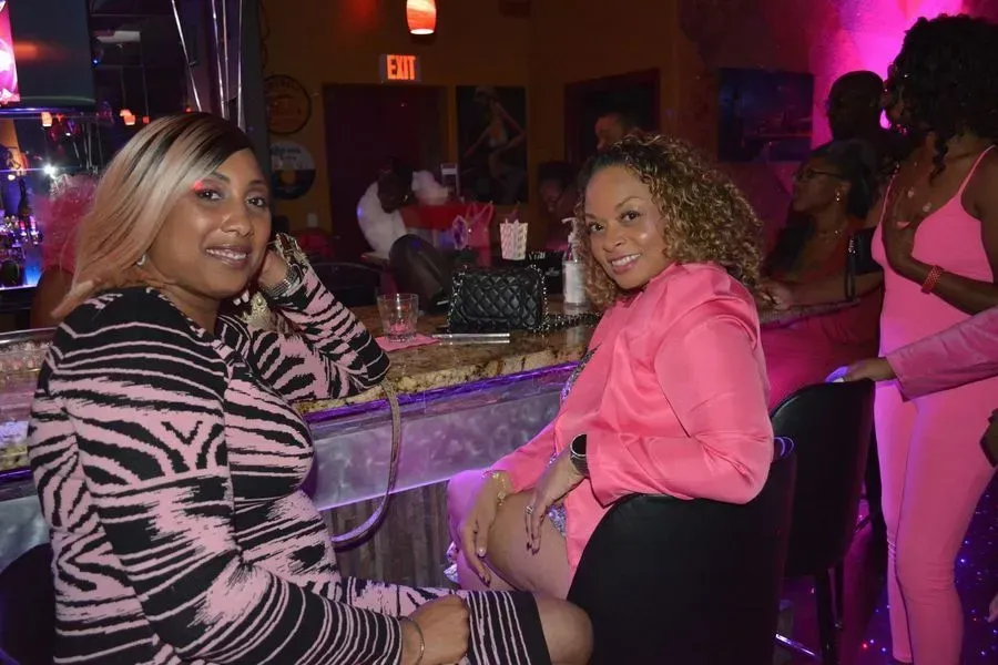 Two women seated at a nightclub bar with pink lighting, facing the camera and smiling