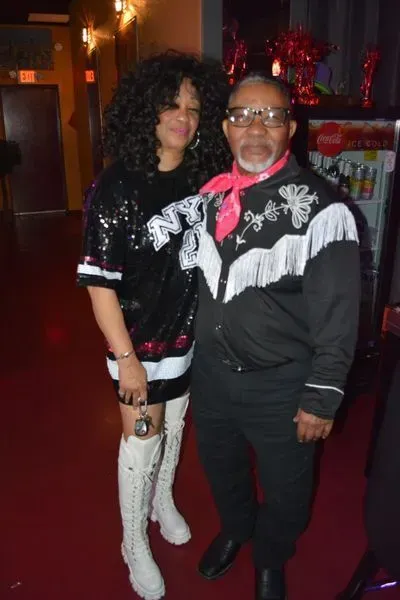Two people posing in a dim nightclub, wearing black and white outfits, smiling at the camera.