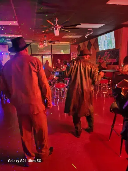 People standing in a red-lit bar, watching a performance on a screen.