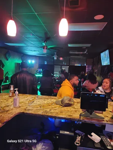 Dimly lit bar with patrons at a granite counter under pink pendant lights and neon glow