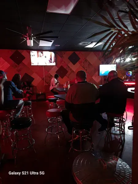 Dim red-lit bar with people seated at tables and stools, ceiling fans, and TVs on the wall.