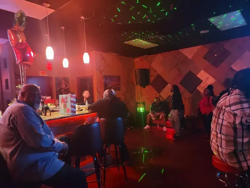 Dimly lit nightclub with a bar, red neon lights, and people socializing on the dance floor.