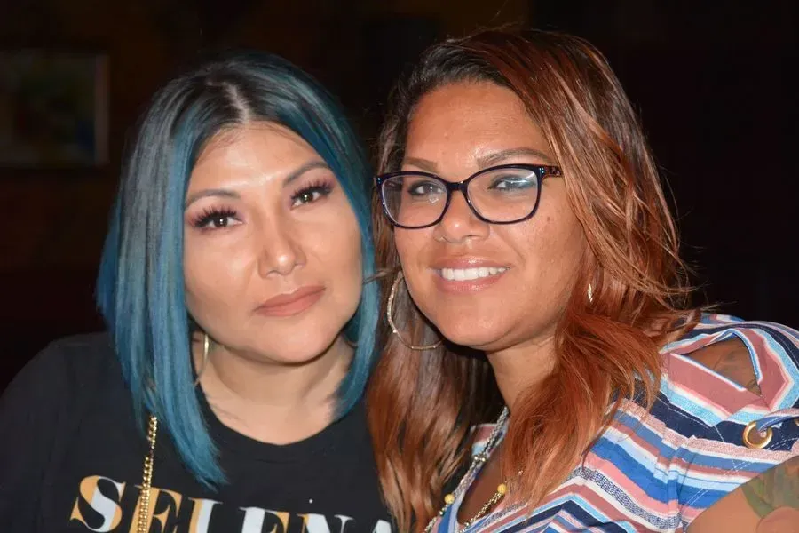 Two women smiling closely together, one with blue hair and one wearing glasses, against a dark background