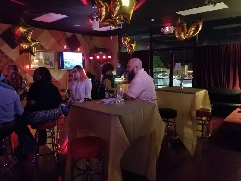 Dim bar/lounge with people chatting around tall tables under red lights and gold balloons