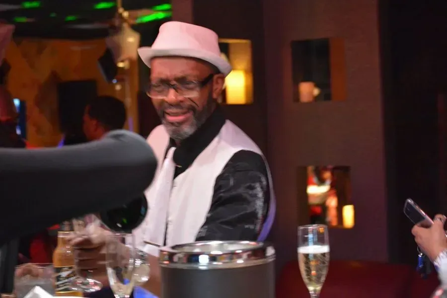 Smiling man in a white hat and vest at a dimly lit event table with drinks and a microphone