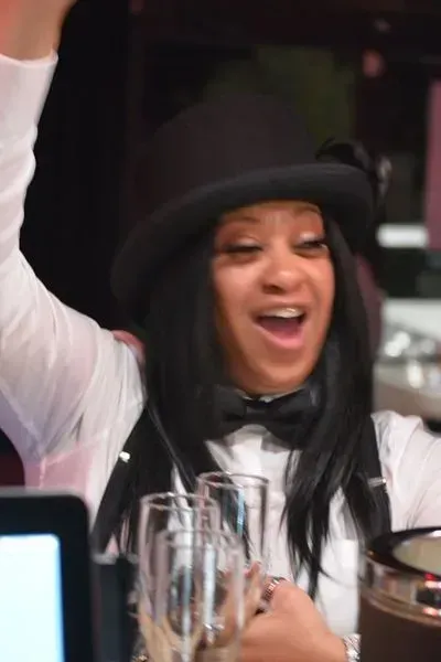 Person in a black hat and vest smiling with one arm raised at a table in a lively indoor setting