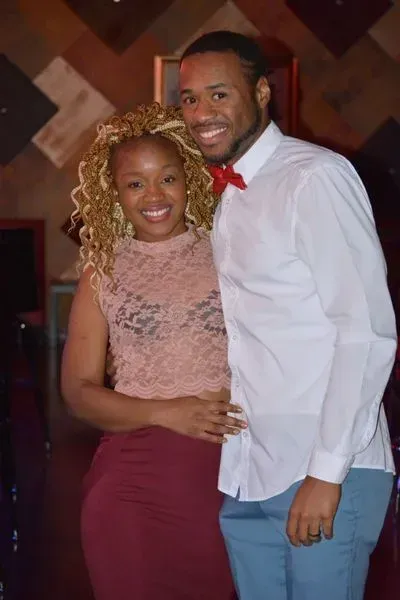 Couple posing together indoors, smiling in formal attire against a patterned backdrop.