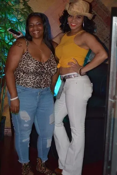 Two women posing together indoors, one in a leopard-print top and jeans, the other in a yellow top and white pants.