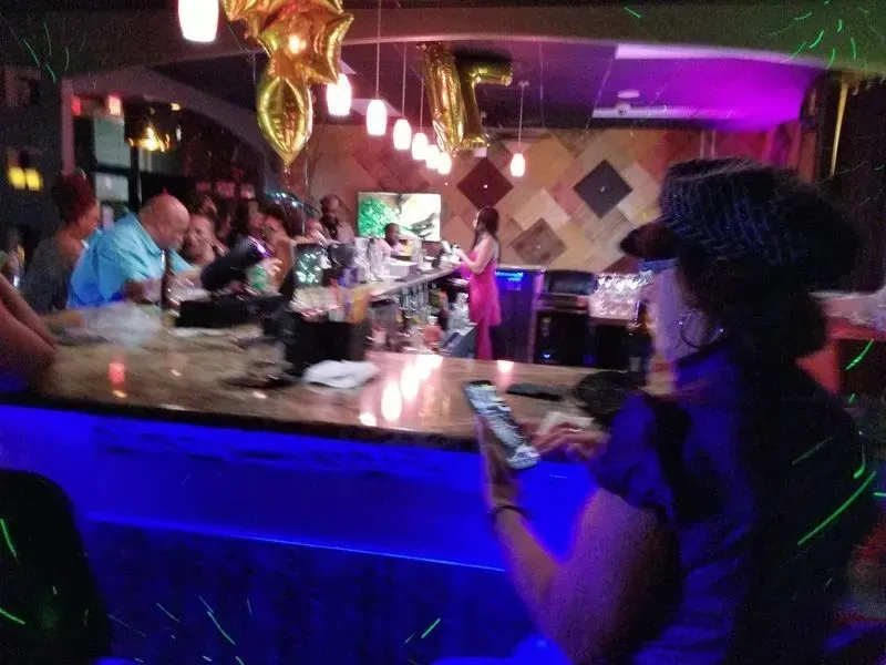 Dimly lit bar with people chatting, blue-lit counter, and a woman using a smartphone in the foreground