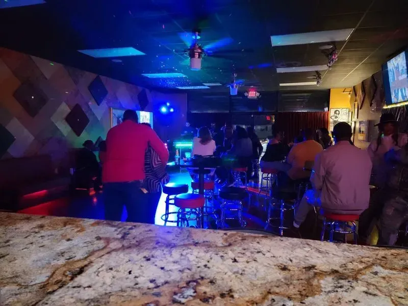 Dim bar interior with people seated at tables under blue and purple lights.