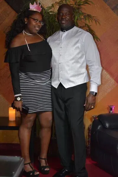 Two people posing indoors, one in a black-and-white dress with a pink crown, the other in a white shirt and black slacks