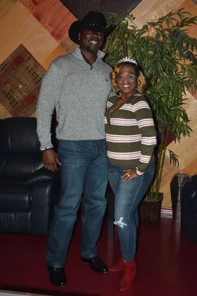 Two people standing indoors, smiling beside a black couch and tall plant.