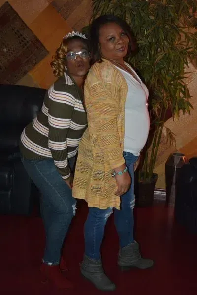 Two people posing indoors, one in a striped sweater and jeans, the other in a tan cardigan and boots.