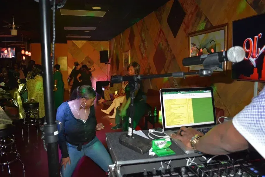 DJ booth in a dim nightclub, with a laptop on the mixer and people dancing in the background