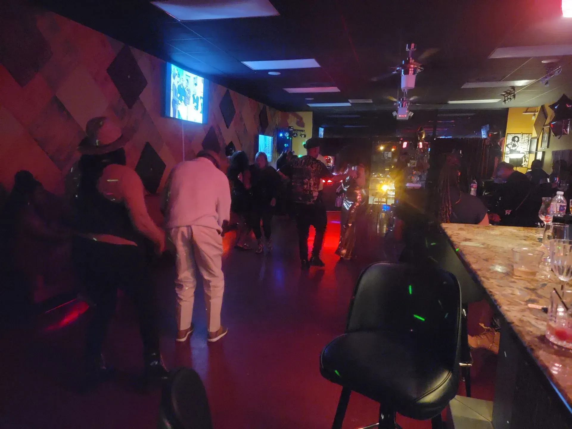 Dim nightclub interior with people standing, colorful lights, and a bar counter on the right.