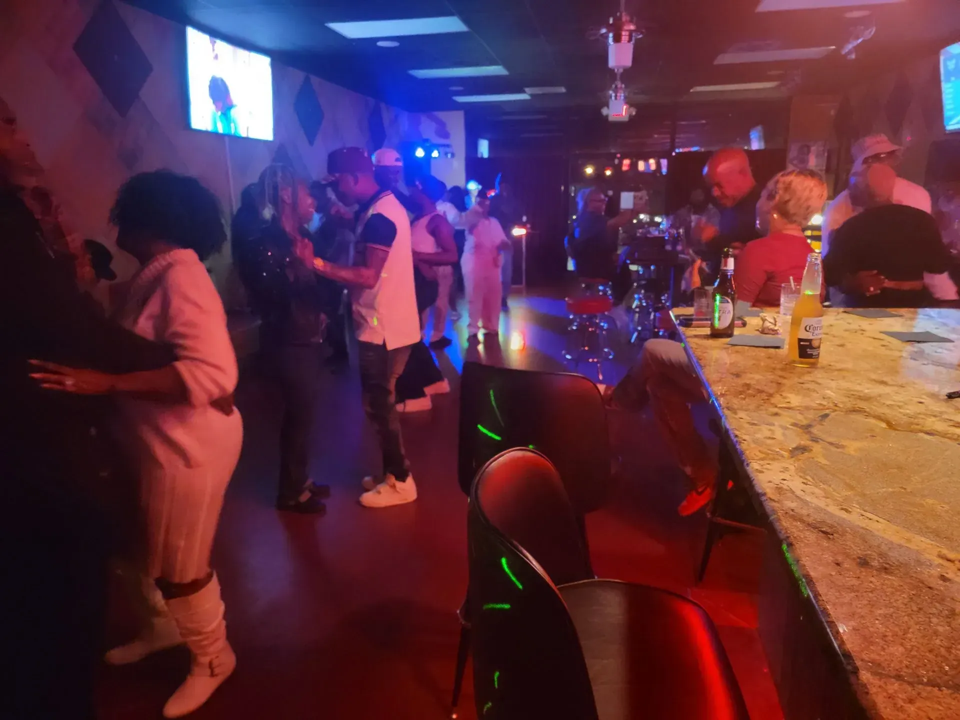 Dimly lit bar with people socializing, red stools, and a lit TV screen in the background.
