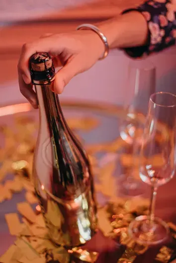 Hand opening a champagne bottle beside two glasses and rose petals under warm pink lighting