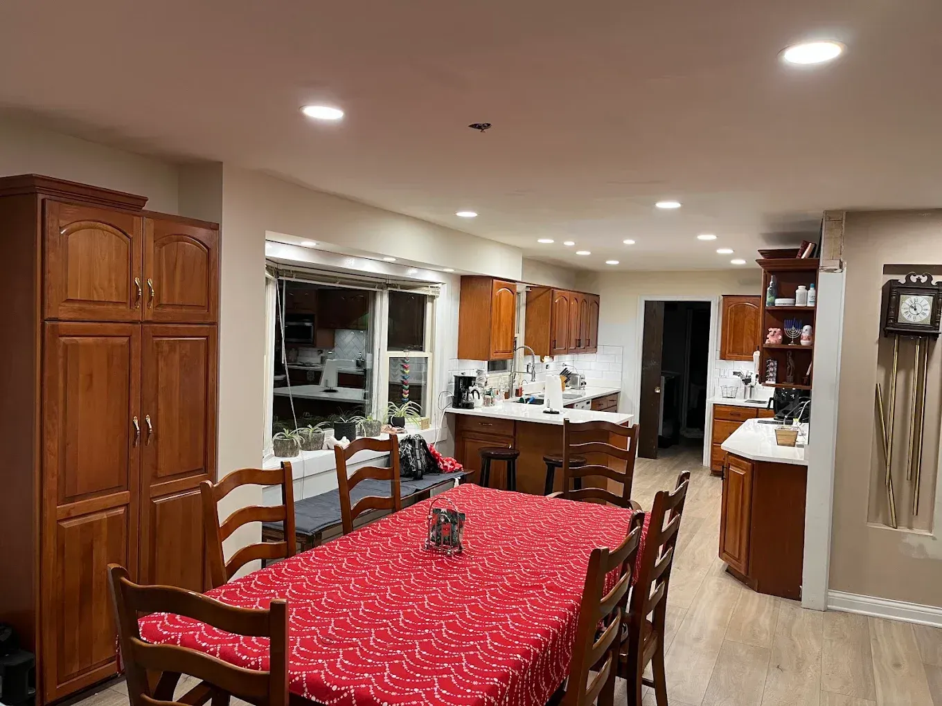 Dining room with a red tablecloth, wooden chairs, and kitchen cabinets under recessed ceiling lights.