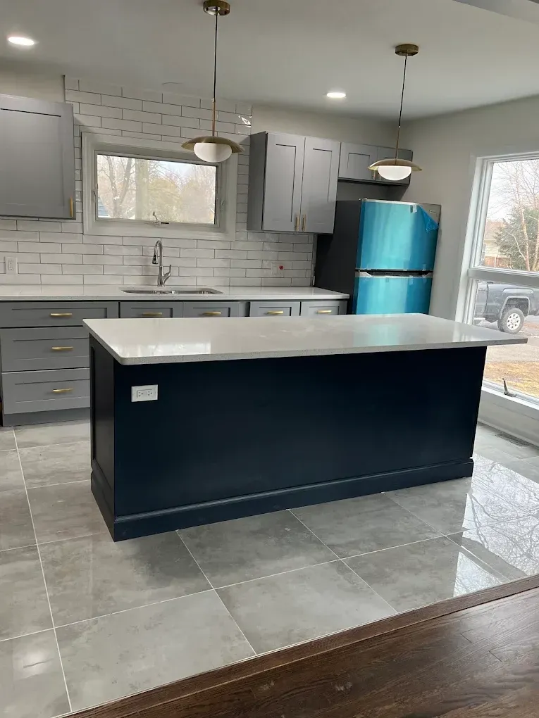 Modern kitchen with gray cabinets, tiled floor, and a dark island; bright blue refrigerator near a window.
