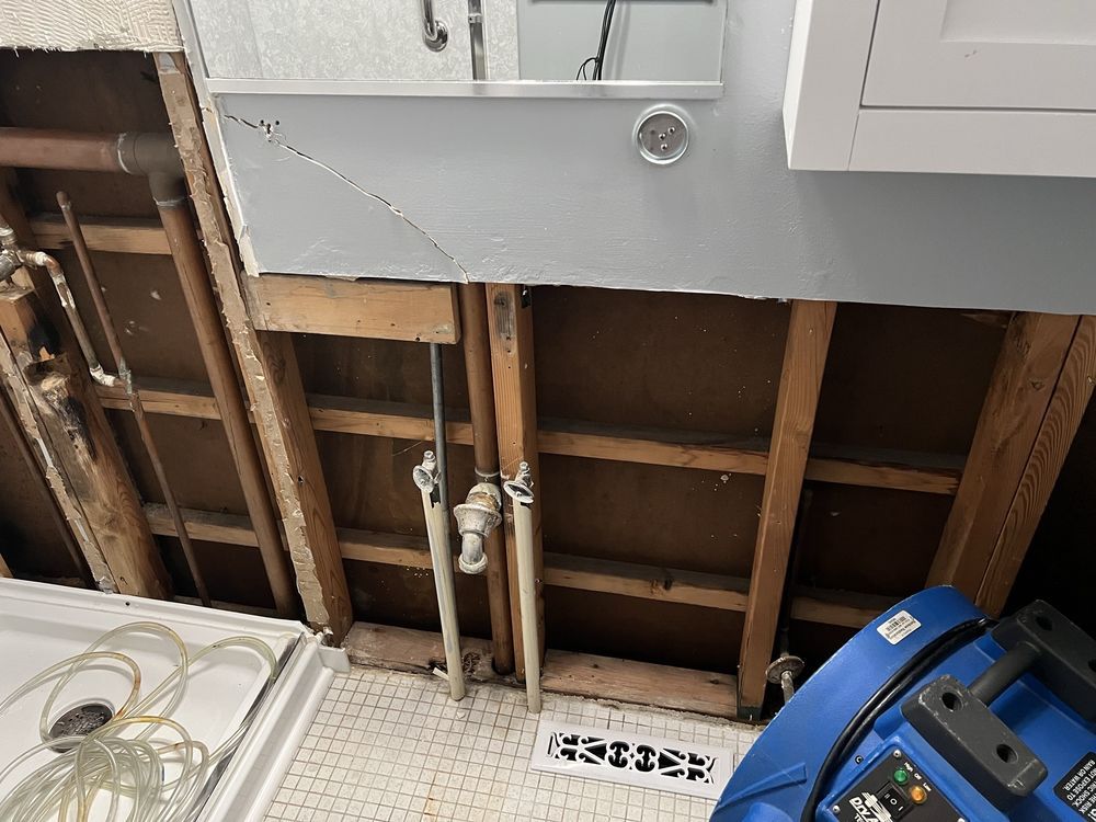 Exposed bathroom wall under sink with plumbing pipes, damaged drywall, and a blue plastic bucket on the floor