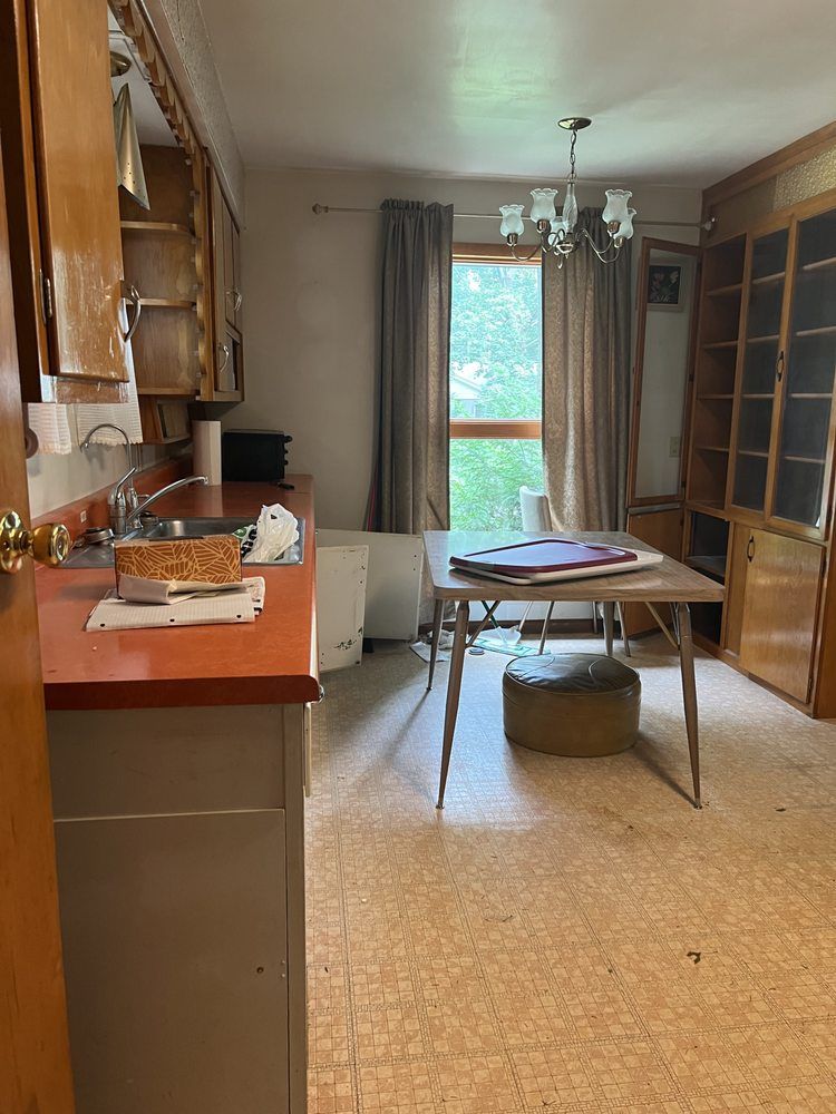 Small kitchen-dining room with orange counters, glass cabinet, round table, and a window with curtains.