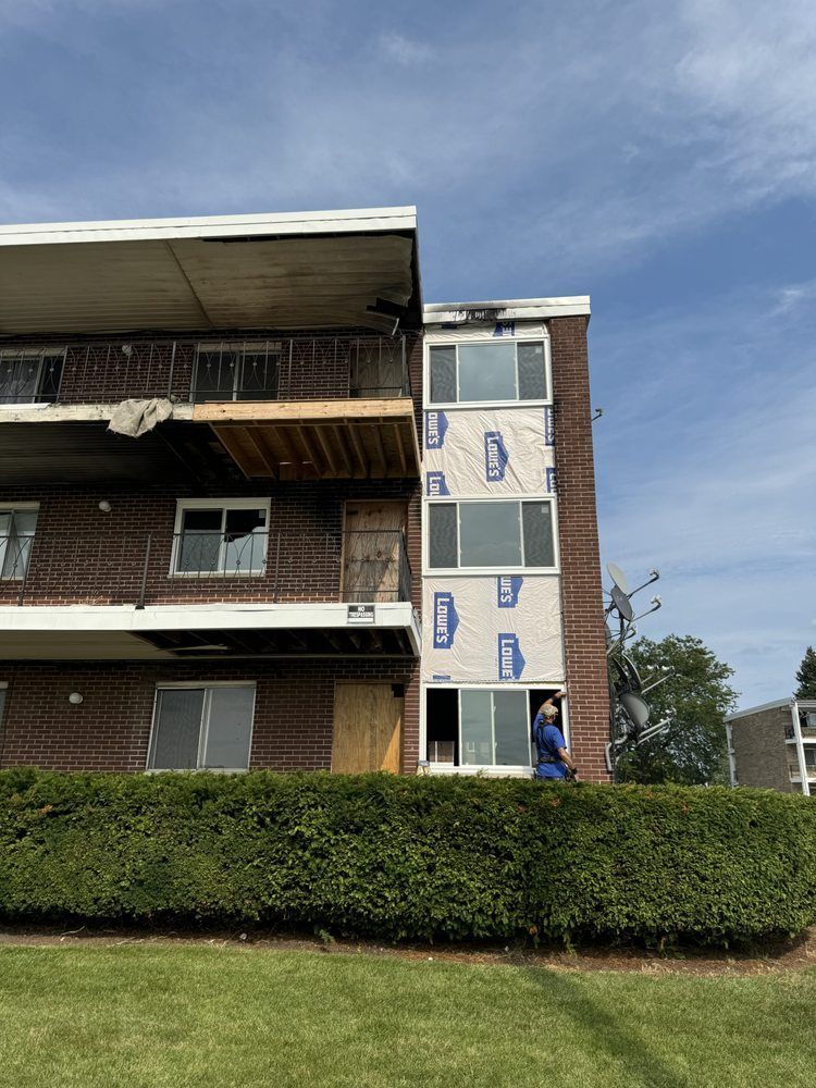 Apartment building with balconies, a white exterior stairwell, and a hedge in front under a cloudy sky