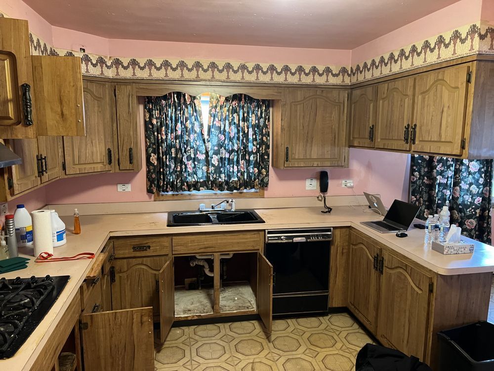 Small vintage kitchen with wood cabinets, pink walls, patterned curtains, and a sink under the window.