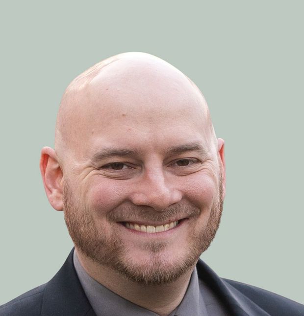 Smiling bald man with a short beard in a dark suit against a plain gray background