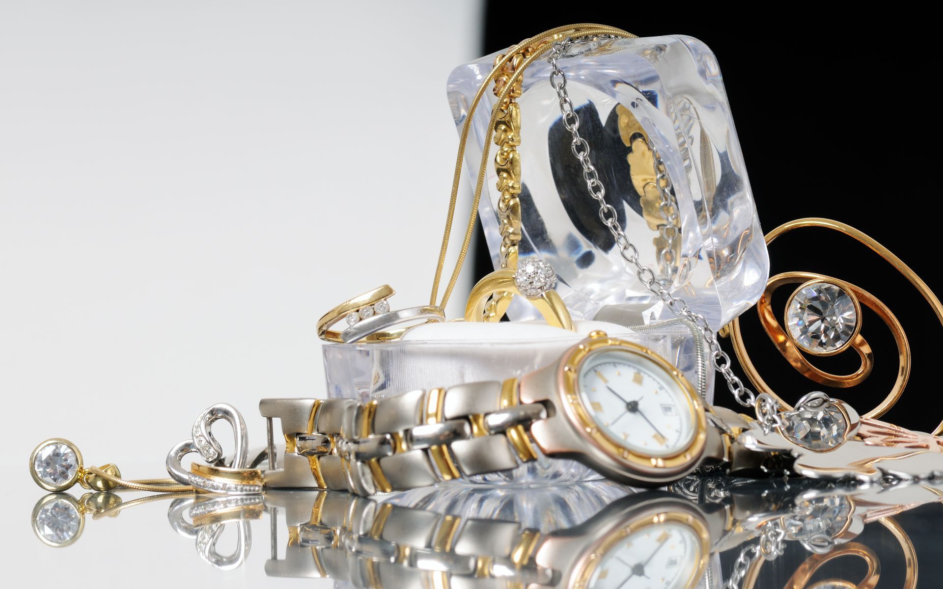 Gold jewelry and watches scattered around a clear cube on a reflective surface