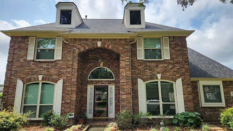 Brick two-story house with arched entryway, shutters, dormers, and dark roof under a cloudy sky.