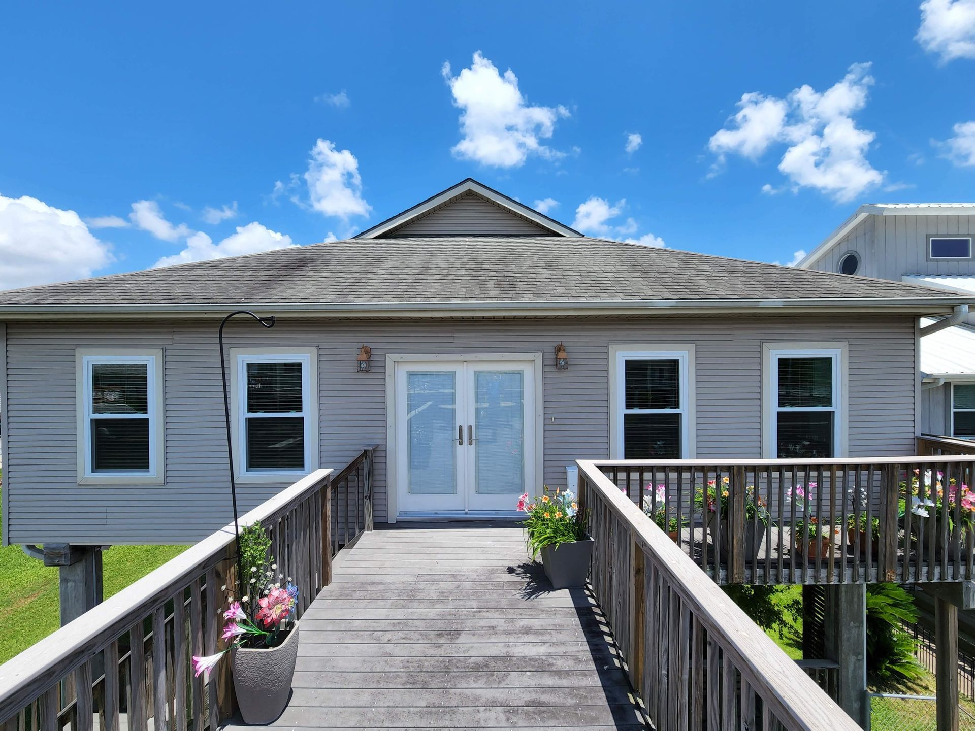 A gray house on stilts with a wooden walkway. White doors, windows, blue sky, and a deck.