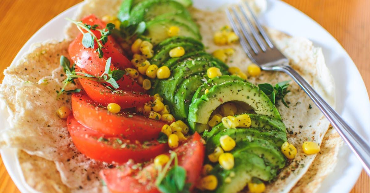 Un plato de comida con aguacate, tomates, maíz y un tenedor sobre una mesa.