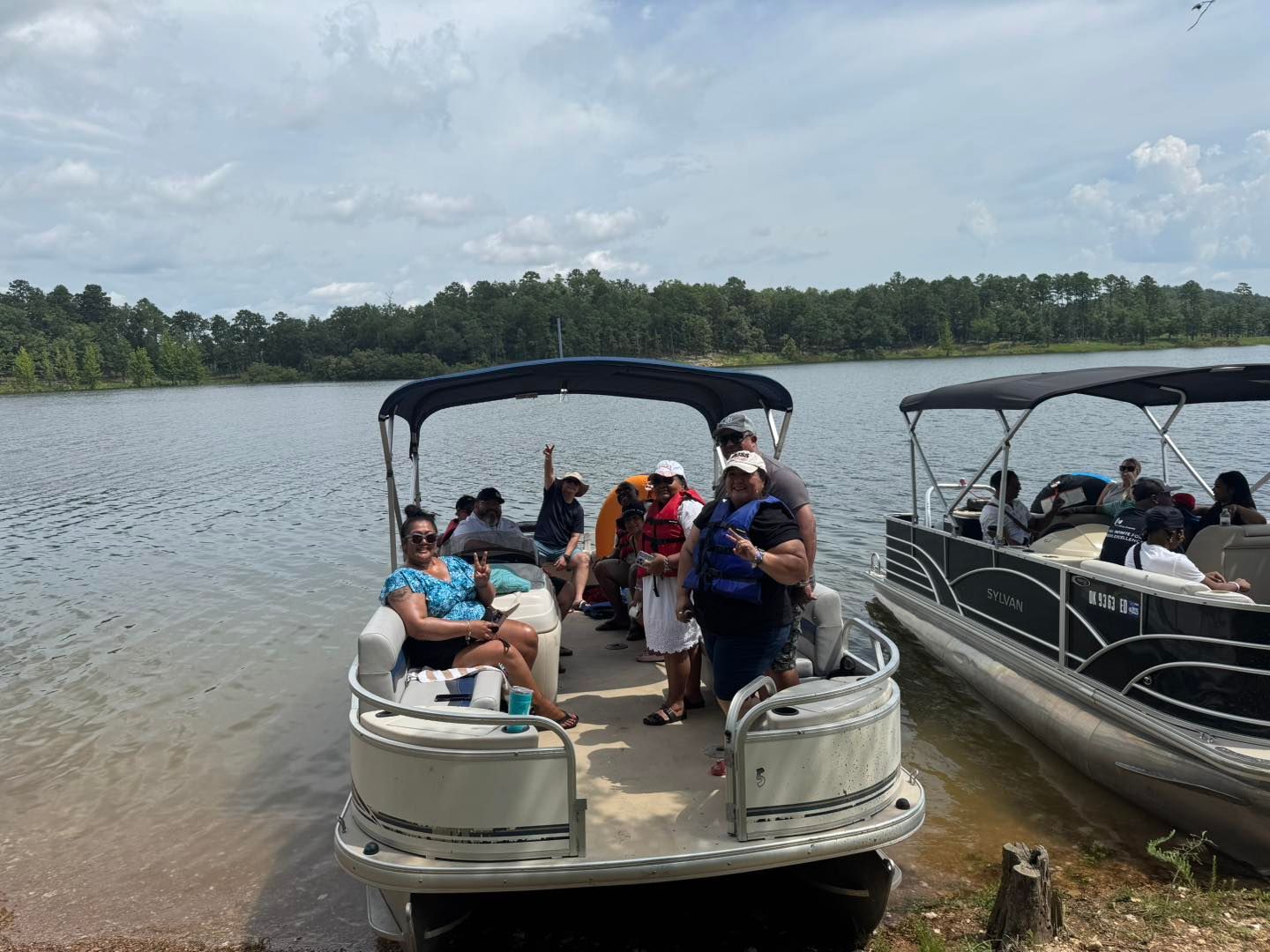 A group of people sitting on a pontoon boat docked at a lake shore, with another boat parked alongside.