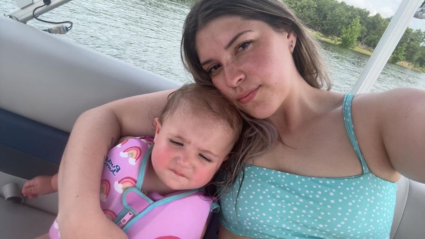 A woman holds a small child in a rainbow-patterned life jacket on a boat, with a lake and trees in the background.