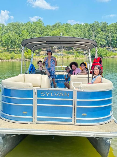 A group of people sitting on a blue Sylvan pontoon boat on a sunny lake with trees in the background.